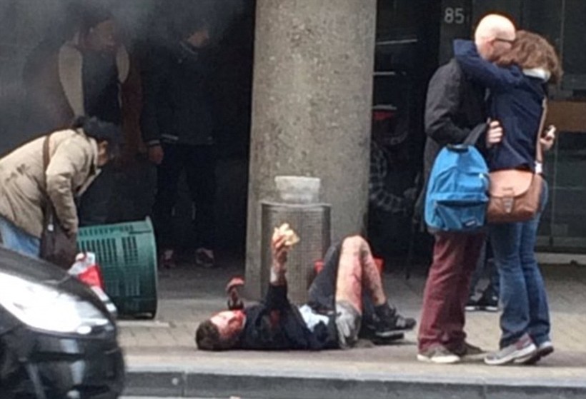 Người đàn ông bị thương đang nằm trên vỉa hè cạnh 2 người vui mừng nhẹ nhõm vì sống sót sau vụ đánh bom tại ga tàu điện ngầm ở Maelbeek
